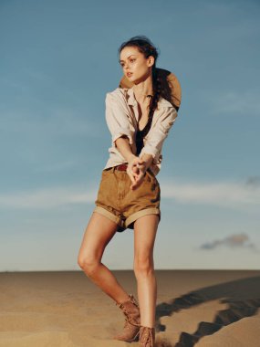 Çölde kumulda duran kadın açık gökyüzünün altında şort ve şapka giyiyor.