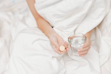 Kadın elinde bir bardak su ve hapla yatakta uzanıyor. Arka planda beyaz bir çarşaf var..