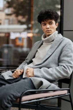 Gri kazaklı ve siyah kotlu bir gencin, pencerenin önündeki sandalyede otururken çekilmiş moda fotoğrafı. Kazağının altına beyaz bir balıkçı yaka giyiyor..