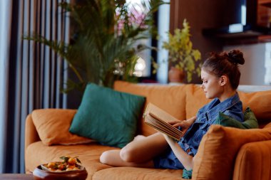 Oturma odasındaki turuncu koltukta oturmuş kitap okuyan ve yemek yiyen rahat bir kadın.