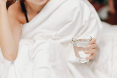 Kadın beyaz yorganın üzerinde bir bardak suyla yatakta oturuyor, kafası yastığın üzerinde dinleniyor..