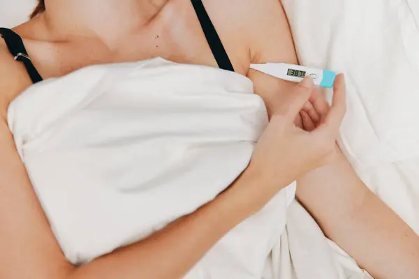 stock image Woman lying in bed holding a thermometer with another thermometer on the bedside table. monitoring body temperature concept.