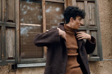 Kahverengi kazak ve ceket giyen, pencerenin önünde tahta panjurlar ve arka planda bir kapı ile poz veren bir kadının moda görüntüsü..