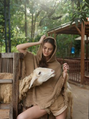 Kahverengi şallı bir kadın bir koyunun yanındaki tahta bankta oturuyor.