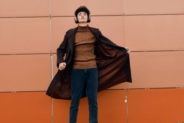 Trençkotlu moda adamı turuncu duvara karşı duruyor elleri ceplerinde ve kafası yana yatık. Moda şehir tarzı konsepti.