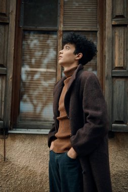 Kahverengi ceketli, siyah balıkçı yaka kazaklı ve pantolonlu şık bir genç, bir pencerenin önünde duruyor ve uzaklığa bakıyor..