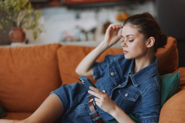 Kot gömlekli kadın kanepede oturmuş oturma odasında cep telefonuna bakıyor.