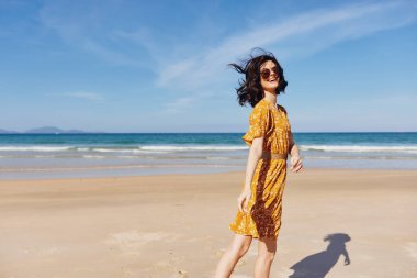 Parlak sarı elbiseli bir kadın güneşli bir günde kumlu plajda engin bir okyanusun arkasında zarif bir şekilde duruyor.