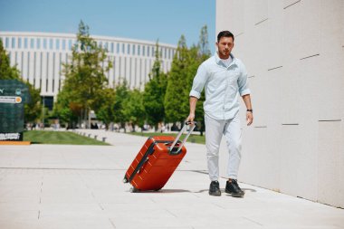 Sıradan giyinmiş genç bir adam şehir parkından kırmızı bir bavul geçirir. Seyahat ya da iş seyahati önerir. Arka planda binalar ve yeşilliklerle modern bir ortam var..
