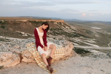 Bir dağın önündeki kayanın tepesinde oturan kadın ekose bir battaniyeyle huzurlu bir yolculuğun tadını çıkarıyor.