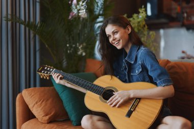 Konforlu ev ortamında pencerenin yanındaki koltukta oturan akustik gitar çalan kadın.
