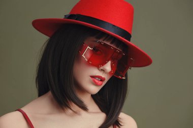 Kırmızı şapkalı, güneş gözlüklü bir kadın şık kırmızı şapkalı ve şık güneş gözlüklü bir kadın fotoğraf için poz veriyor, eli kalçasında güvenle dururken diğeri nazikçe yüzüne dokunuyor, zarafet ve cazibeyle.