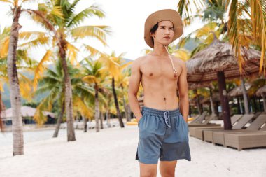 Gülümseyen Asyalı Adam Kaslı Gövde, Şapka ve Yaz Gökyüzü ile Tropikal Plaj Tatilinin keyfini çıkarıyor