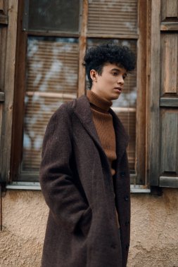 Kahverengi ceketli ve balıkçı yaka kazaklı şık bir genç arka planda ahşap kapılı bir pencerenin önünde dikiliyor..