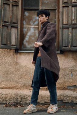 Kahverengi paltolu ve spor ayakkabılı şık bir genç adam eski bir binanın önünde elleri ceplerinde duruyor..