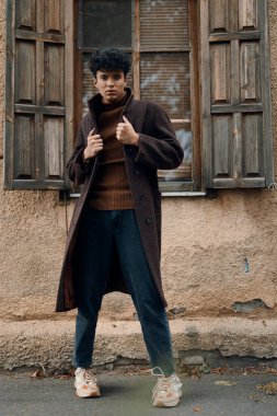 Kahverengi ceketli şık bir adam arka planda penceresi ve önünde kapısı olan klasik bir binanın önünde poz veriyor..