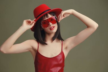 Kırmızı şapkalı ve güneş gözlüklü Asyalı kadın gri arka planda kendine güvenerek poz veriyor, şık bir moda sergiliyor ve zarafetini sergiliyor.