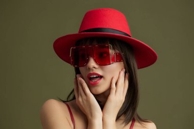 Kırmızı şapka, güneş gözlüğü, kırmızı dudak rengi kırmızı şapka ve güneş gözlüğü takan bir kadın kameraya poz veriyor, elleri yüzünü çerçeveliyor, kırmızı dudak rengini şık ve kendinden emin bir şekilde vurguluyor.