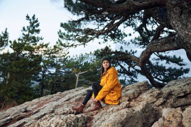 Sarı paltolu bir kadın doğa güzelliğinin tadını çıkarıyor. Seyahat macerası sırasında bir ağacın önündeki bir kayanın üzerinde oturuyor.