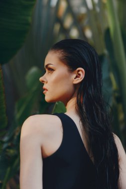 Uzun siyah saçlı güzel bir kadın yemyeşil bir bitkinin önünde duruyor. Arka planda palmiye ağacı var.