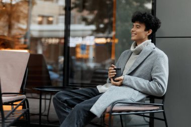 Camın kenarındaki sandalyede oturan, elinde bir fincan kahve tutan ve gülümseyen şık genç adam..