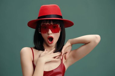 Şok olmuş kırmızı şapkalı ve güneş gözlüklü kadın, elleri başının üstünde, ağzı açık, kameraya bakıyor.