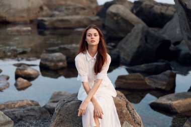 Beyaz elbiseli, dingin bir kadın okyanus kayalığında oturuyor.