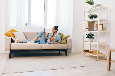 Mutlu bir kadın rahat bir koltukta oturuyor, akıllı bir telefon tutuyor ve gülümsüyor online oyunlardan zevk alıyor ve güzel oturma odasında mesajlar okuyor..