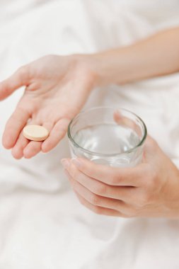 Elinde bir bardak su ve beyaz çarşaflı ve arka planda yastıklı bir hap tutan bir kadın. Uyumadan önce ilaç alma kavramı..