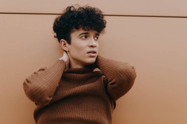 Kahverengi balıkçı yaka kazaklı kıvırcık saçlı adam elleri başının üzerinde duvara yaslanıyor. Modaya uygun bir erkek model..