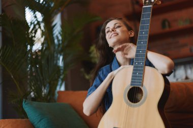 Konforlu oturma odasındaki çiçeğin önündeki koltukta gitar çalan kadın.