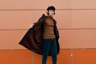 Kahverengi kazaklı ve ceketli genç adam turuncu duvarın önünde elleri ceplerinde ve kulaklıkları kulağında duruyor. moda konsepti.
