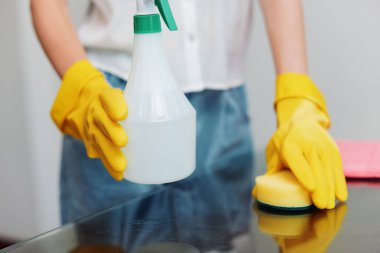 Sarı lastik eldivenli bir kadın temizlik malzemesi tutuyor ve sarı eldivenli beyaz gömlekli başka bir kadının önünde cam masaya sünger sürüyor. İki kadın birlikte temizlik yapıyor..