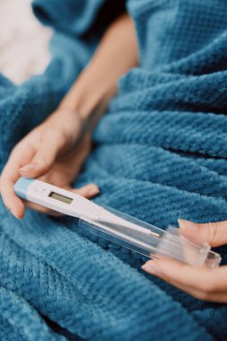 Woman holding thermometer in bed with blue blanket on head, another thermometer in hand. clipart