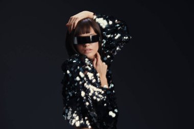Sequin ceket, güneş gözlüğü pullu ceket ve güneş gözlüklü bir kadın siyah arka planda poz veriyor, elleri başının üstünde, başı yana eğik, büyüleyici, kendinden emin bir ruh hali yakalıyor.