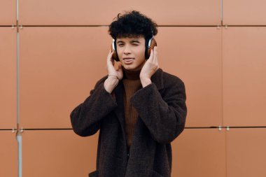 Kafasında kulaklıkla müzik dinleyen, turuncu bir duvarın önünde duran kahverengi ceketli şık bir genç..