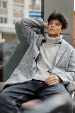 Gri ceketli, beyaz balıkçı yaka kazaklı, elleri başının üzerinde bankta oturan şık bir genç..