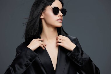 Siyah takım elbiseli ve güneş gözlüklü bir kadın, gri bir arka planda duruyor, elleri kalçasında rahatça poz veriyor.