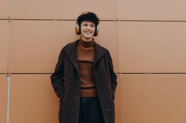 Turuncu duvara karşı duran kulaklıklı bir gencin moda portresi. Kahverengi bir ceket ve siyah bir balıkçı yaka giyiyor..