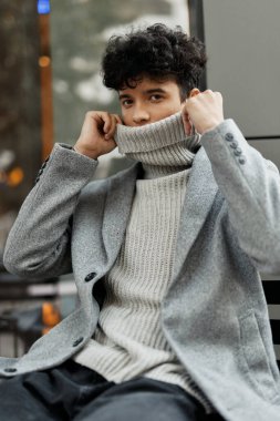 Moda adamı elleri yüzün üzerinde bankta oturuyor balıkçı yaka kazak, kazak ve ceket giyiyor..