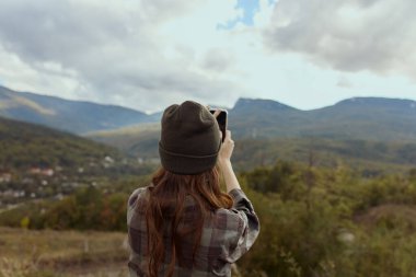 Şık şapkalı bir kadın seyahat macerası sırasında kamerayla görkemli bir dağ manzarası çekiyor.