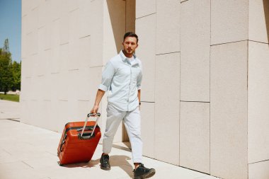 Güncel bir binanın dışında turuncu bir bavulla yürüyen kendine güvenen genç adam seyahat, iş yeri ya da taşınma öneriyor..