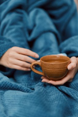 Mavi battaniye ve yorgana sarılı bir fincan çay tutan kadın..