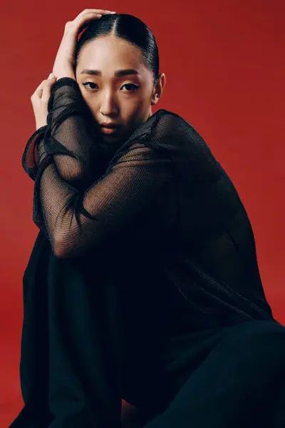 stock image Asian fashion: a woman in a sheer top and black pants sitting on a red background with her hands on her face and eyes closed.