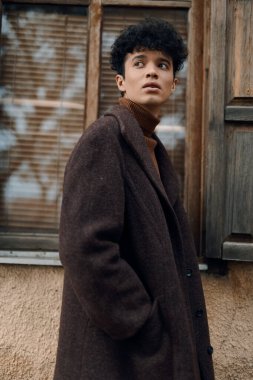 Kahverengi paltolu şık bir genç arka planda bir ağaçla pencerenin önünde kameradan başka yöne bakıyor..