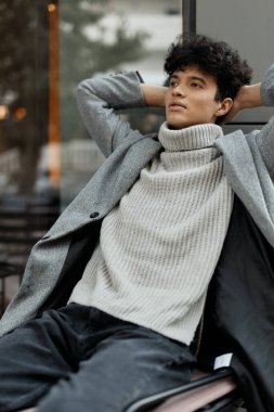 Gri kazaklı ve elleri başının arkasında siyah pantolonlu bir adam bankta oturuyor..