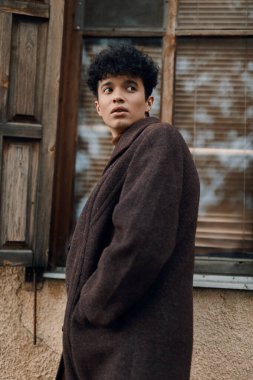 Kahverengi paltolu şık genç adam eski binanın önünde duruyor. Arka planda ahşap kapı ve açık pencere var..