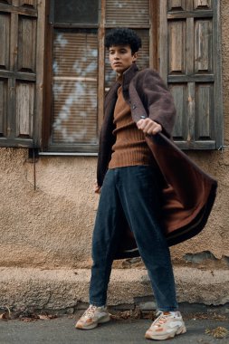 Eski bir binanın önünde kahverengi ceketli, kazaklı ve mavi pantolonlu şık bir adam duruyor. Görünüşünü beyaz spor ayakkabılarla tamamlıyor..