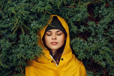 Sarı yağmurluklu genç bir kadın. Etrafı hep yeşil ağaçlar ve çalılarla çevrili. Huzurlu bir ortam.