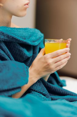 Mavi battaniyeli kadın elinde bir bardak portakal suyuyla yatakta oturuyor. kafasının altında yastık.
